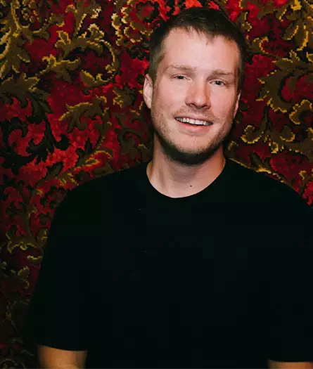 A man in a black shirt smiling in front of an ornate wallpaper.